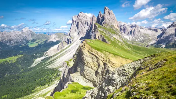 Dramatic view of the Dolomites in Selva, with jagged mountain peaks towering over alpine meadows and forested slopes under a clear sky.