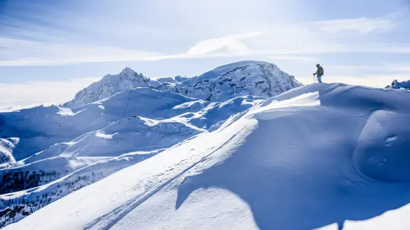 A lone skier stands atop a majestic French mountain at sunrise, surrounded by snow-capped peaks and a sea of clouds, basking in the serene beauty of the Alps.